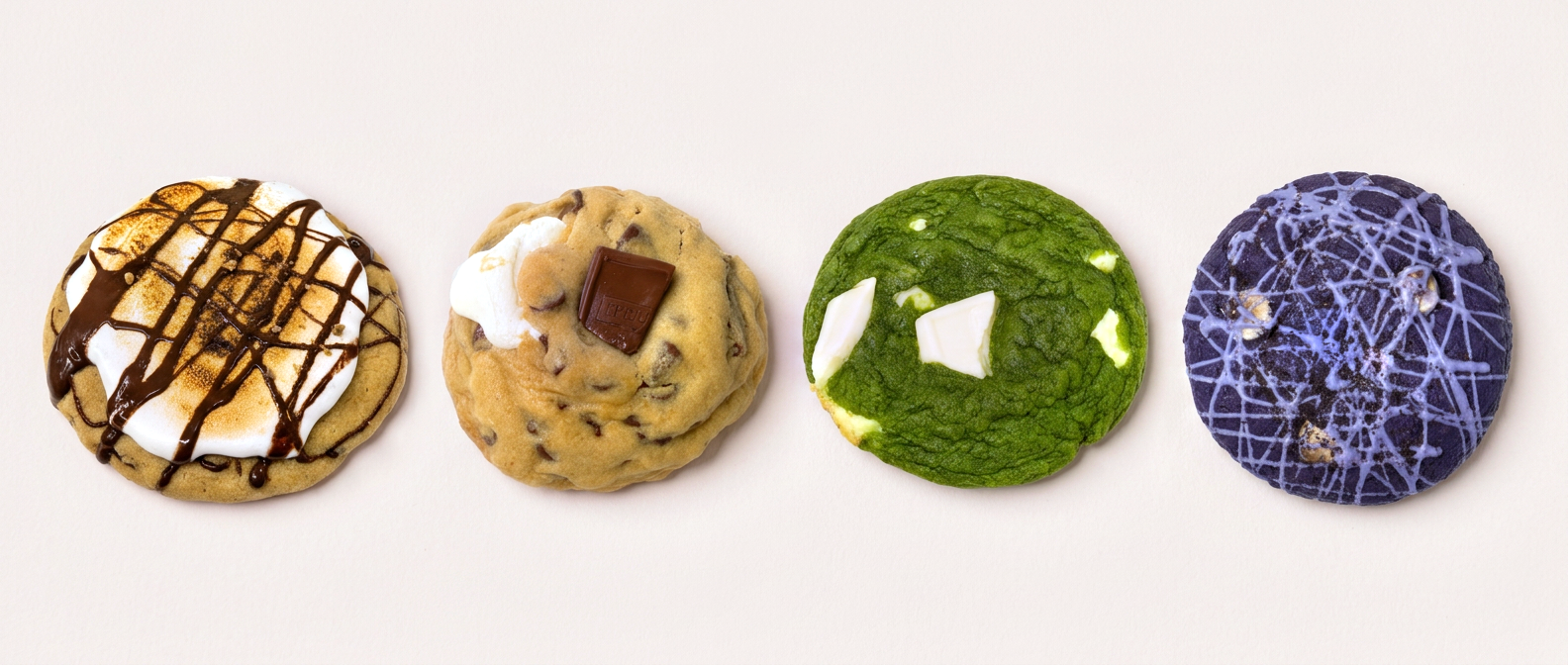 Four cookies in a row on a blush surface: toasted marshmallow with chocolate drizzle, a chocolate chunk cookie with an embedded chocolate square, matcha with white chocolate chunks, and a purple cookie with lavender drizzle